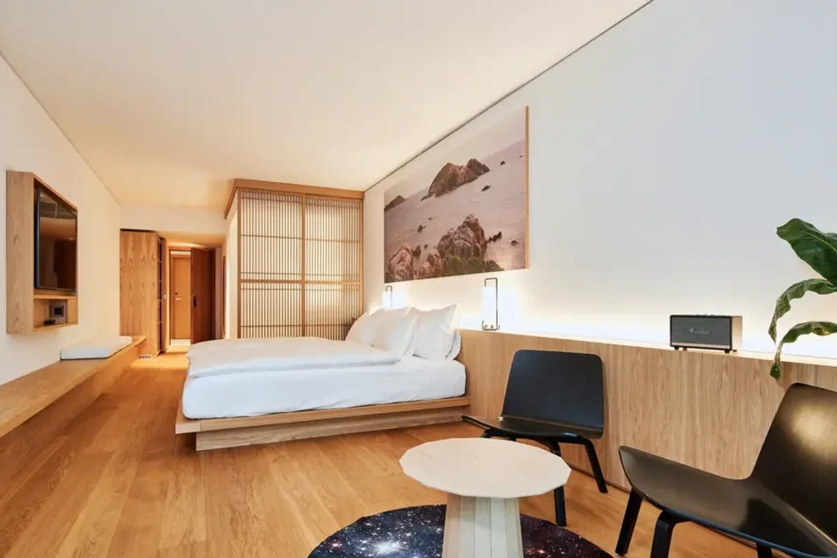 Aydınlık ahşap zemini, alçak yatağı, Japon esintili paravanı ve yatağın üzerindeki kayalık sahil tablosuyla modern bir otel odası; minimalist siyah sandalyeler ve galaksi desenli halı, Basel'deki tasarım odaklı konaklama seçeneklerini yansıtan Japon estetiğini ve çağdaş sanatı bir araya getiriyor.