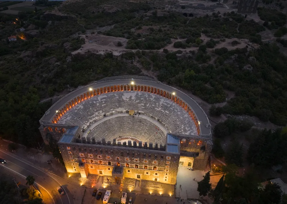 Aspendos Örenyeri'ndeki Antik Tiyatro'nun, koyu renkli tepelerle çevrili loş bir gece vaktinde, taş basamaklı oturma alanları ve görkemli sahne binasının sıcak sarı ışıklarla aydınlatıldığı, aşağıda hareketli bir yol ve giriş alanını gösteren büyüleyici bir havadan görünüm.