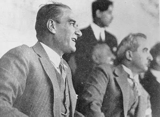 Mustafa Kemal Atatürk, Gazi Koşusu'nun coşkulu atmosferinde, çizgili kravatlı ve şık bir takım elbiseyle profilden neşeyle gülümsüyor; bu tarihi siyah beyaz karede arkasında diğer davetliler de görülüyor.