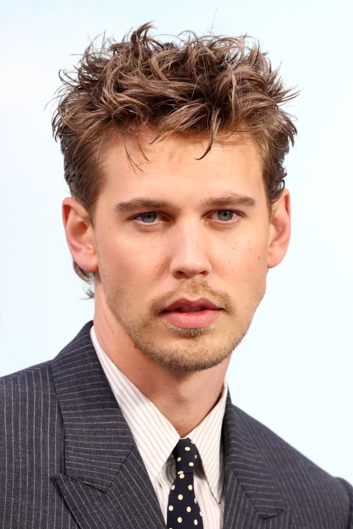 Austin Butler, The Bikeriders galasında açık kahverengi dağınık saçları, hafif bıyığı ve sakalıyla, koyu çizgili takım elbisesi, ince çizgili gömleği ve puantiyeli kravatıyla objektife doğrudan bakarak poz veriyor.