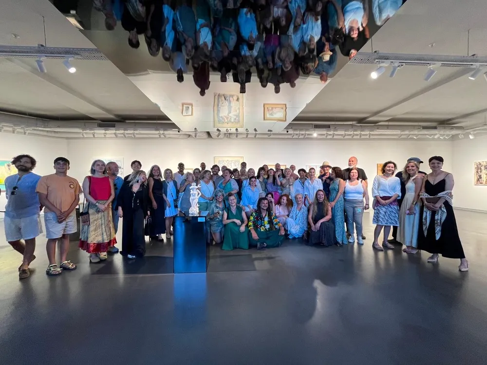 Parlak tavanında ters yansımaları görünen bir sanat galerisinde, duvarlarda eserlerin ve ortada bir heykelin bulunduğu ortamda, kalabalık bir sanatçı grubu fotoğraf çekimi için bir araya gelmiş neşeyle poz veriyor.