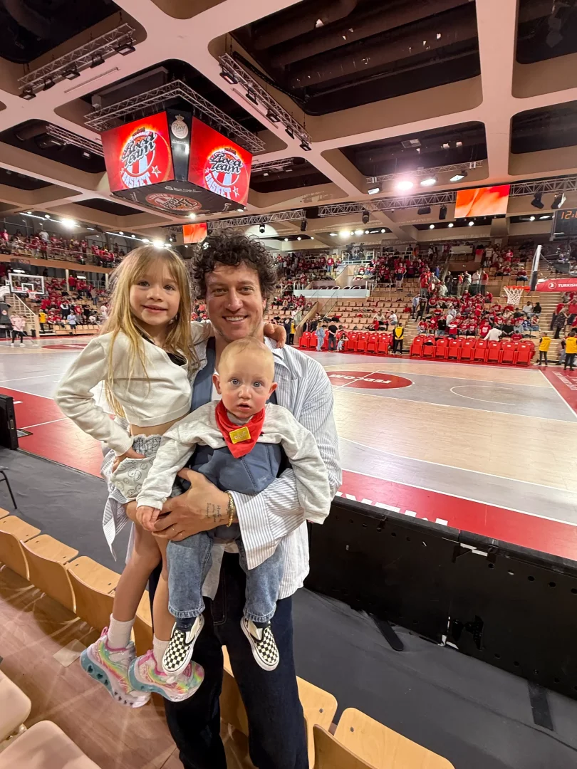 Babalık konusundaki Babalar Günü yazısı bağlamında, sporcu Caner Erdeniz, kalabalık bir basketbol sahasında kendisi gibi gülümseyen uzun sarı saçlı kızı ve kucağındaki kırmızı bandana takmış minik bebeğiyle poz vererek, çocuklarıyla kurduğu derin ve neşeli bağı gözler önüne seriyor.