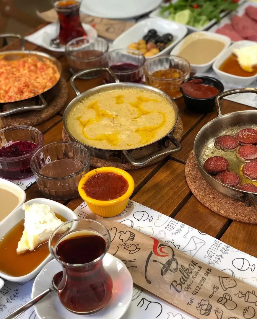 Ahşap bir masanın üzerinde, BalKon Cafe'ye özgü serpme kahvaltının zenginliği gözler önüne seriliyor; sıcak menemen, eritilmiş tereyağlı kuymak, kızarmış sucuk, çeşitli reçeller, bal kaymak, zeytinler ve demli bir çay bardağıyla dolu bu enfes Türk kahvaltısı tüm detaylarıyla iştah açıyor.