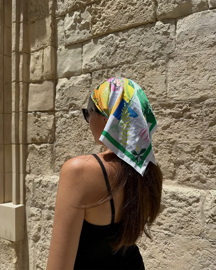 Güneşin vurduğu taş duvara karşı sırtı dönük duran, kahverengi saçlarına tropikal desenli rengarenk bir bandana bağlamış kadın, bandanalı saç modellerine modern ve şık bir yorum katıyor.