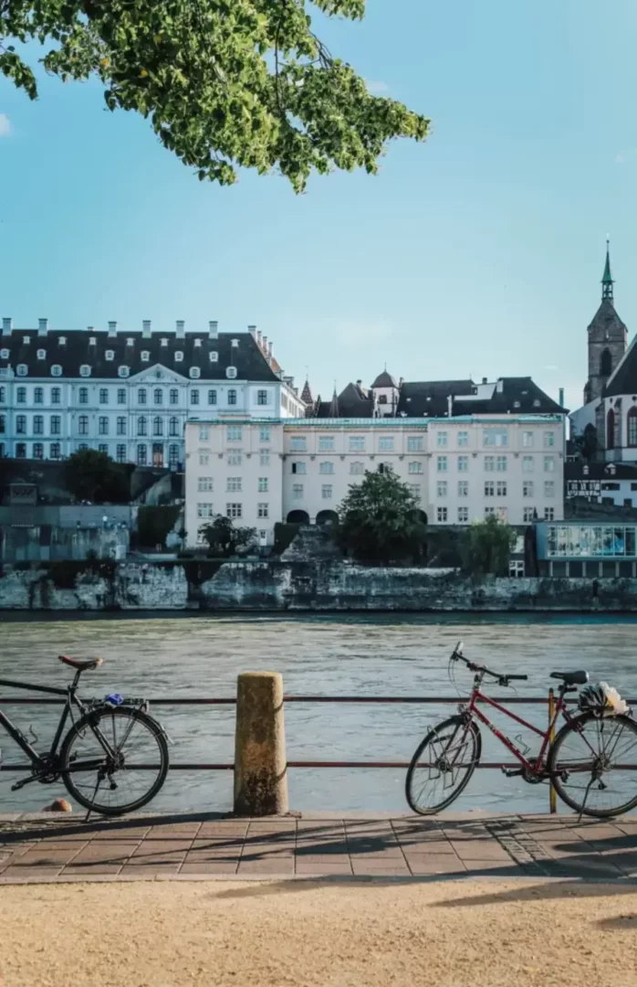Basel gezilecek yerler rotasında Ren Nehri kıyısında tarihi binalar, kilise kulesi ve bisikletlerle görünen güneşli şehir manzarası.