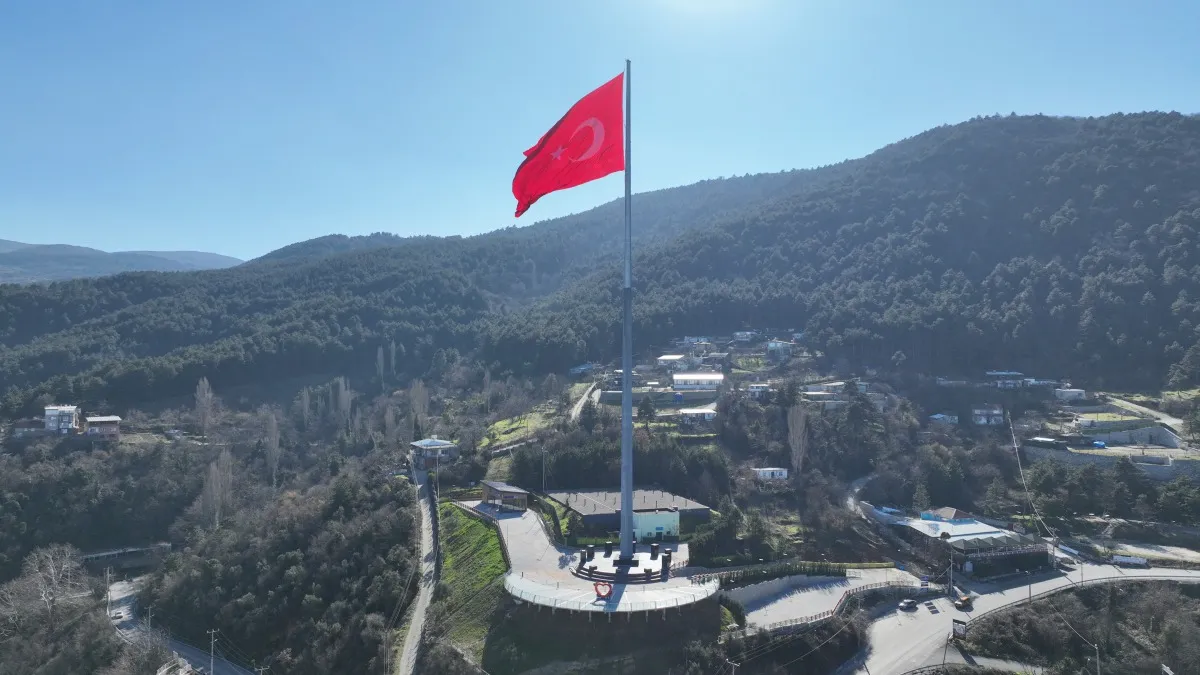 Yemyeşil tepelerin ve masmavi gökyüzünün ortasında gururla dalgalanan devasa bir Türk bayrağı, Bayraktepe'deki yüksek direğinde duruyor, etrafında virajlı yollar ve dağınık yerleşimler bulunan bu görkemli manzara, bölgenin simgesel bir noktasını gözler önüne seriyor.