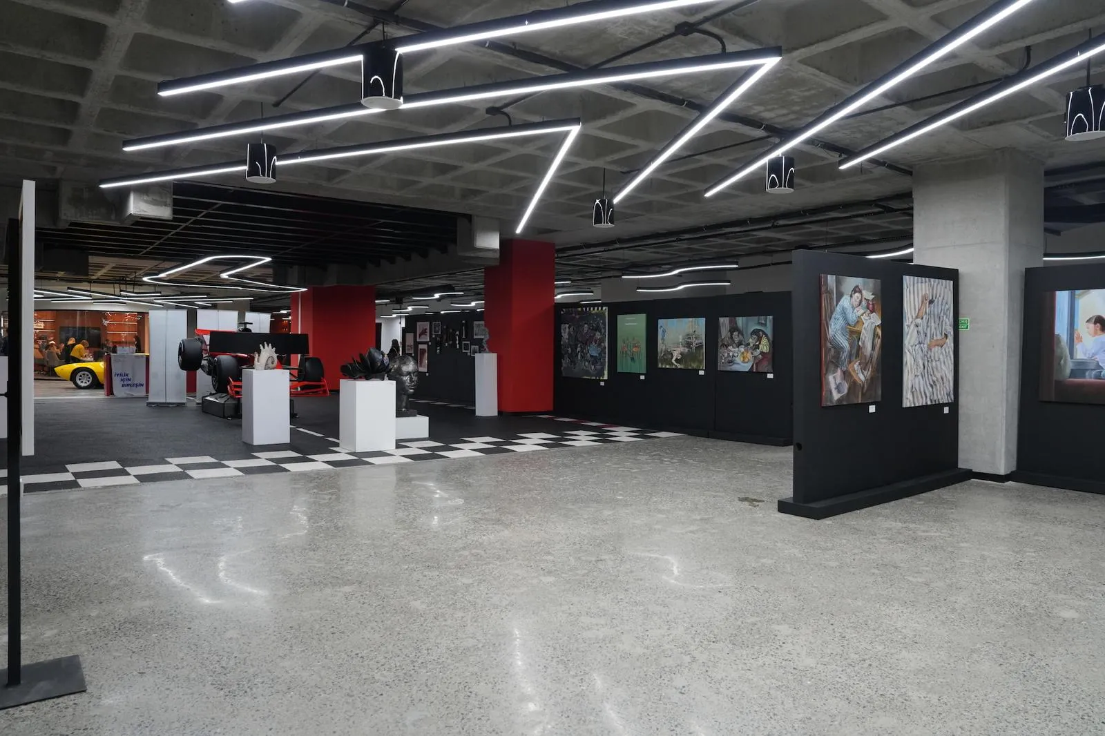 Modern ve geniş bir sanat fuarı veya bazaart sergi alanında, siyah panellerdeki çok sayıda tablonun ve beyaz kaidelerdeki heykellerin yanı sıra arka planda parlak sarı bir klasik otomobil ile yarış arabası parçalarının da yer aldığı, mimari ışıklandırmasıyla dikkat çeken, hareketli bir görüntü.