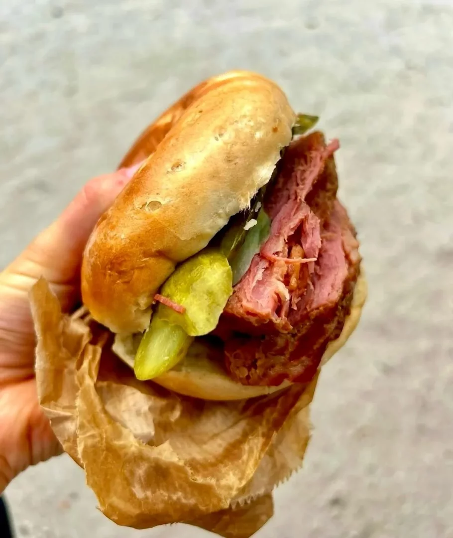 Bir elin kahverengi kağıda sararak tuttuğu, taze fırınlanmış altın sarısı bagel ekmeği arasında bolca dilimlenmiş pembe füme et ve parlak yeşil salatalık turşuları ile hazırlanmış iştah açıcı bir bedigel sandviç.