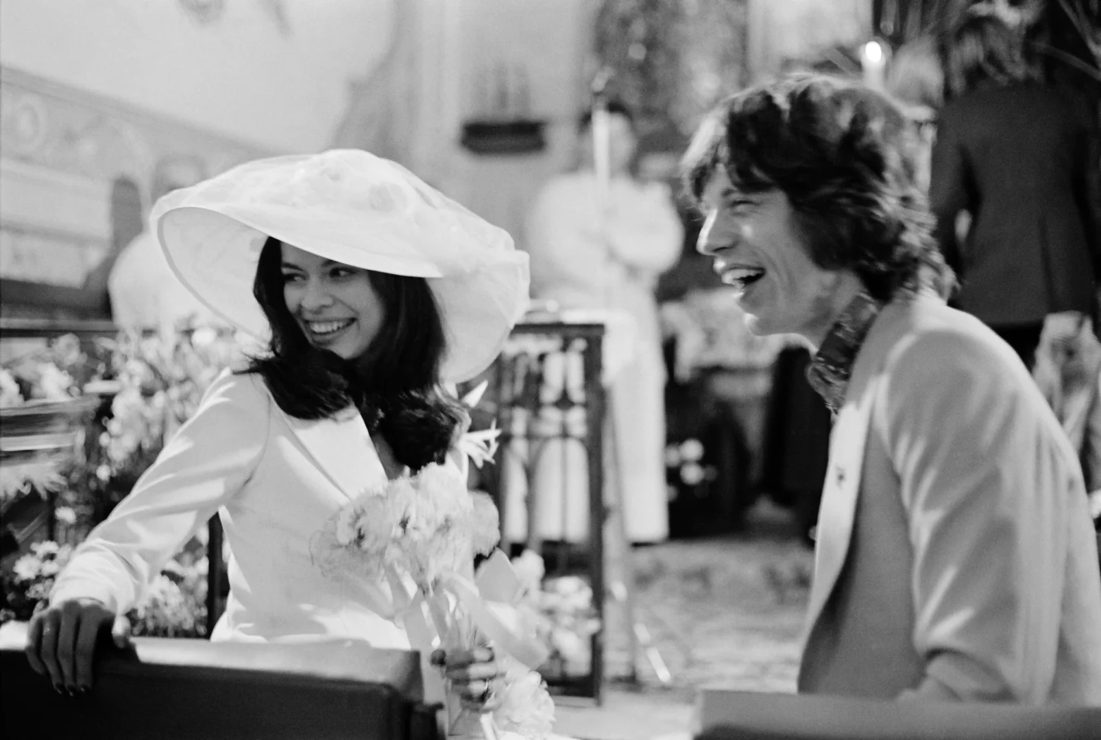 Siyah beyaz fotoğrafta, beyaz geniş kenarlı şapkası ve blazer ceketiyle ikonik gelinlik stilini yansıtan Bianca Jagger, elinde çiçek buketiyle neşeyle gülümserken, sağında ona doğru dönük, kahkahalarla gülen Mick Jagger ile düğün gününden samimi bir an paylaşıyor.
