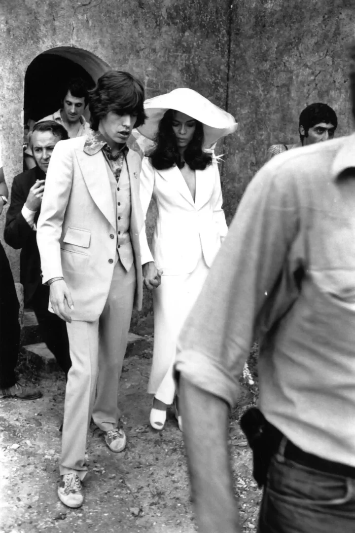 Moda tarihinin ikonik anlarından biri olan bu siyah beyaz fotoğrafta, Bianca Jagger ve Mick Jagger düğün günlerinde el ele yürürken, Bianca'nın derin V yakalı beyaz blazer, uzun etek ve geniş kenarlı şapka kombini zamansız gelinlik anlayışına radikal bir duruş sergiliyor.