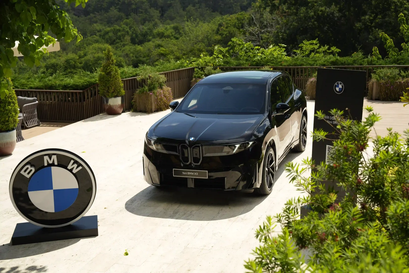 Siyah renkli Yeni BMW iX3 elektrikli SUV, güneşli bir açık hava etkinliğinde, büyük bir BMW logosu ve bilgilendirme paneli eşliğinde, yeşil bitki örtüsüyle çevrili şık bir terasta sergilenerek dikkat çekiyor.