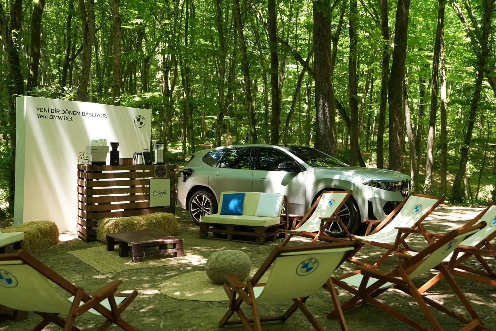 Ormanlık bir alanda, bir ağacın yanında şarj edilen gümüş rengi Yeni BMW iX3 elektrikli SUV, etrafında ahşap paletlerden yapılmış bir kafe, hasır balyaları ve BMW logolu şezlonglarla donatılmış, doğayla iç içe bir tanıtım alanında sergileniyor.