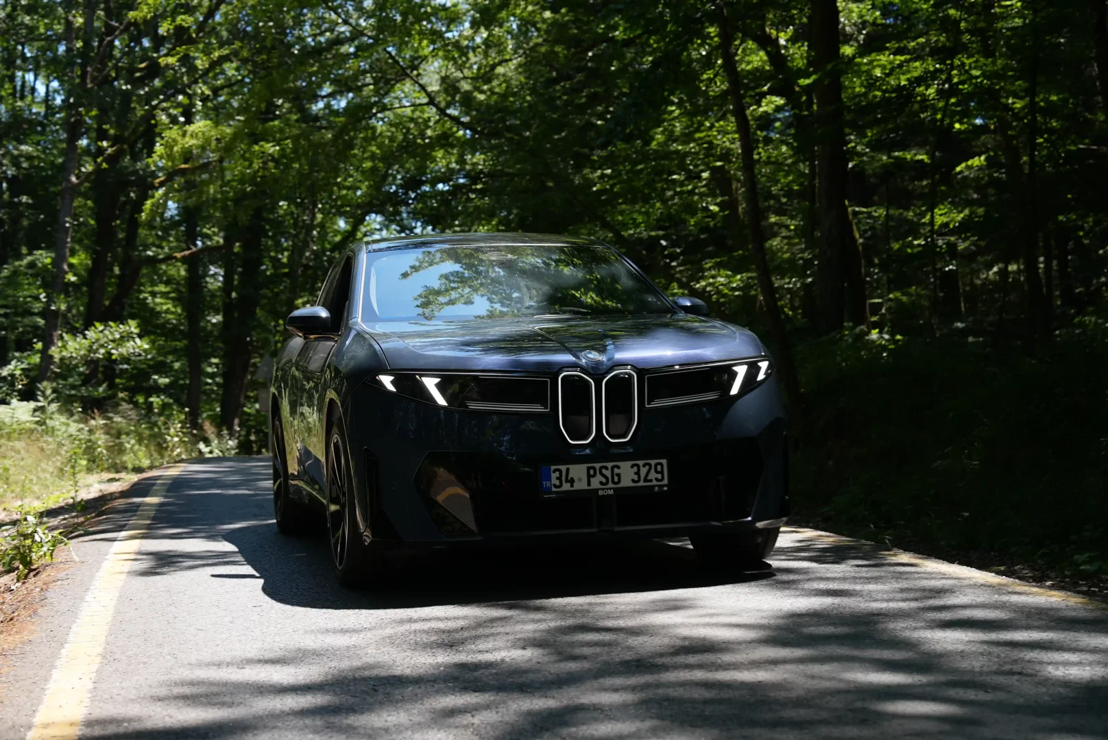 Koyu renkli, modern hatlara sahip bir BMW yeni model sedan, ormanlık alandaki virajlı asfalt yolda, güneş ışınlarının ağaç yaprakları arasından süzülerek yolu aydınlattığı bir anında ilerliyor; öndeki özel tasarımlı aydınlatmalı ızgarası ve keskin LED farları belirgin.
