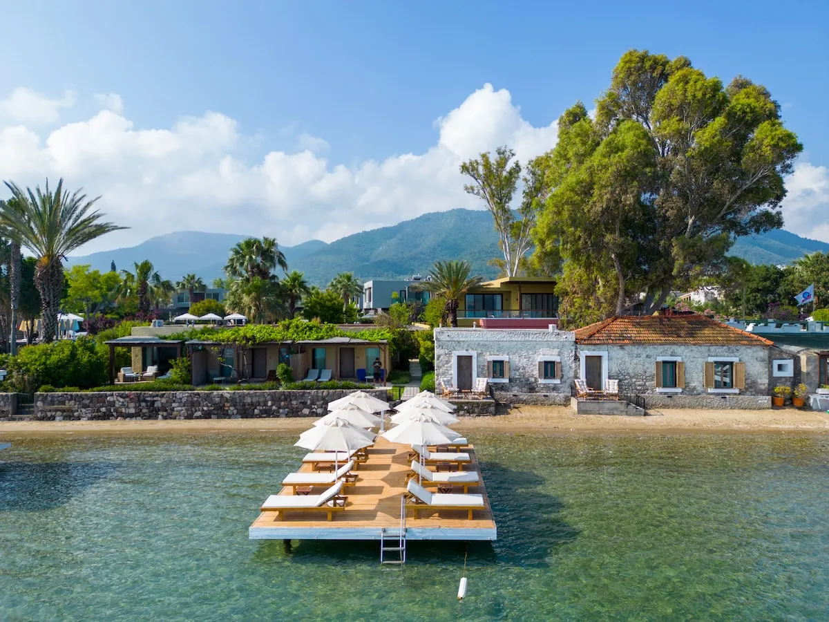 Bodrum Flamm'ın berrak turkuaz sularına uzanan ahşap iskelede beyaz şemsiyeler ve şezlonglar sıralanırken, arkasında kıyıya yakın iki geleneksel taş ev, daha geride modern otel binaları ve yemyeşil ağaçlarla kaplı dağ manzaralı huzurlu bir sahil manzarası.