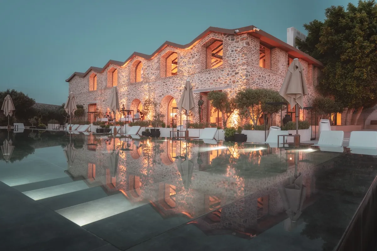 Bodrum'da tarihi dokulu taş mimarisiyle dikkat çeken Ruins Luxury Resort, kemerli pencerelerinden sızan sıcak ışıklarla aydınlanırken, terasındaki misafirler ve şezlonglu havuzunun sakin suyunda görkemli yansımalarıyla huzurlu bir akşam atmosferi sunuyor.