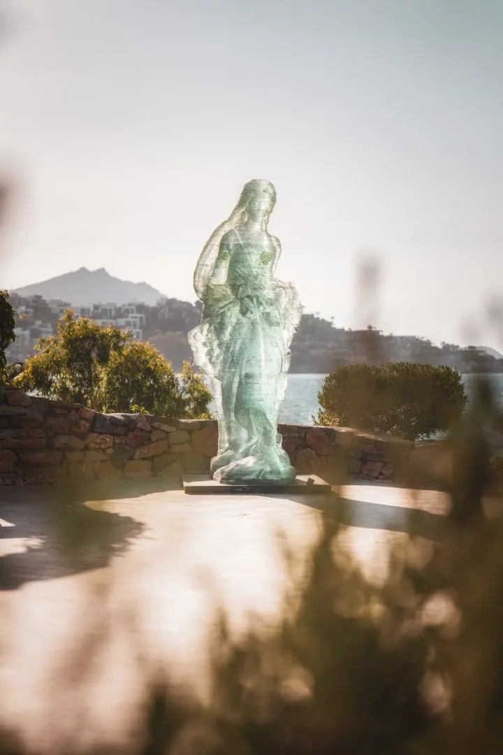 Bodrum'daki Ruins Luxury Resort deniz manzaralı terasında, arka planda Ege Denizi ve tepelerle çevrili, katmanlı camdan yapılmış gibi görünen, şeffaf, kadın figürlü modern bir heykel duruyor.