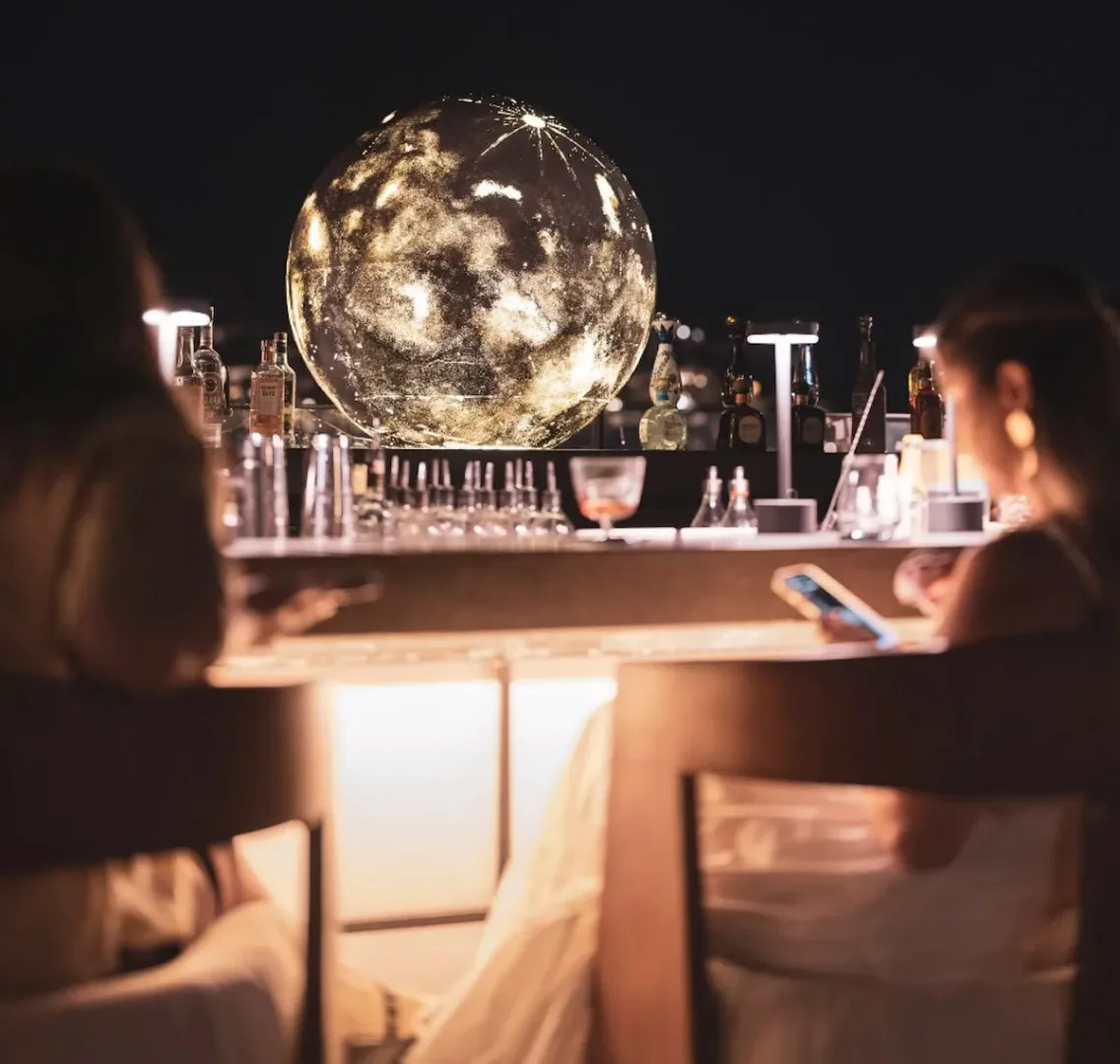 Bodrum'un göz alıcı bir barında, iki kişi parlak ay benzeri devasa bir kürenin aydınlattığı modern bir ortamda kokteyllerini yudumlarken, etraftaki şişeler ve kadehler sofistike atmosferi tamamlıyor.