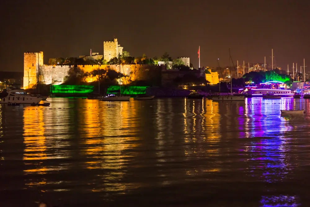 Bodrum Kalesi'nin geceki görkemli manzarasını sergileyen bu fotoğrafta, kalenin altın sarısı ve yeşil ışıklarla aydınlatılan surları ile Sualtı Müzesi'nin bulunduğu alan, denizde yüzen tekneler ve renkli yansımalar eşliğinde göz kamaştırıyor.