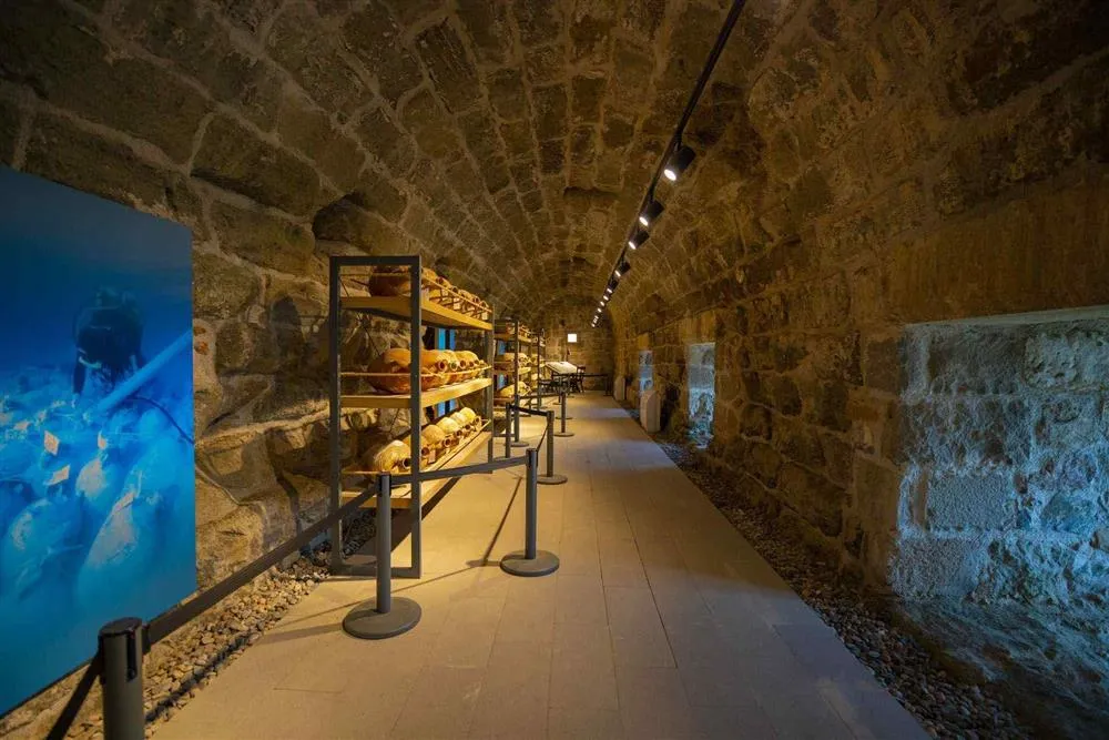 Bodrum Sualtı Müzesi'nin tonozlu taş duvarlı bir galerisinde, sol taraftaki dalgıç temalı büyük mavi sergi panosu ve önündeki raflarda dikkatlice düzenlenmiş çok sayıda amfora benzeri antik eser sergileniyor, koridor boyunca modern aydınlatma ve bariyerler görülebilir.