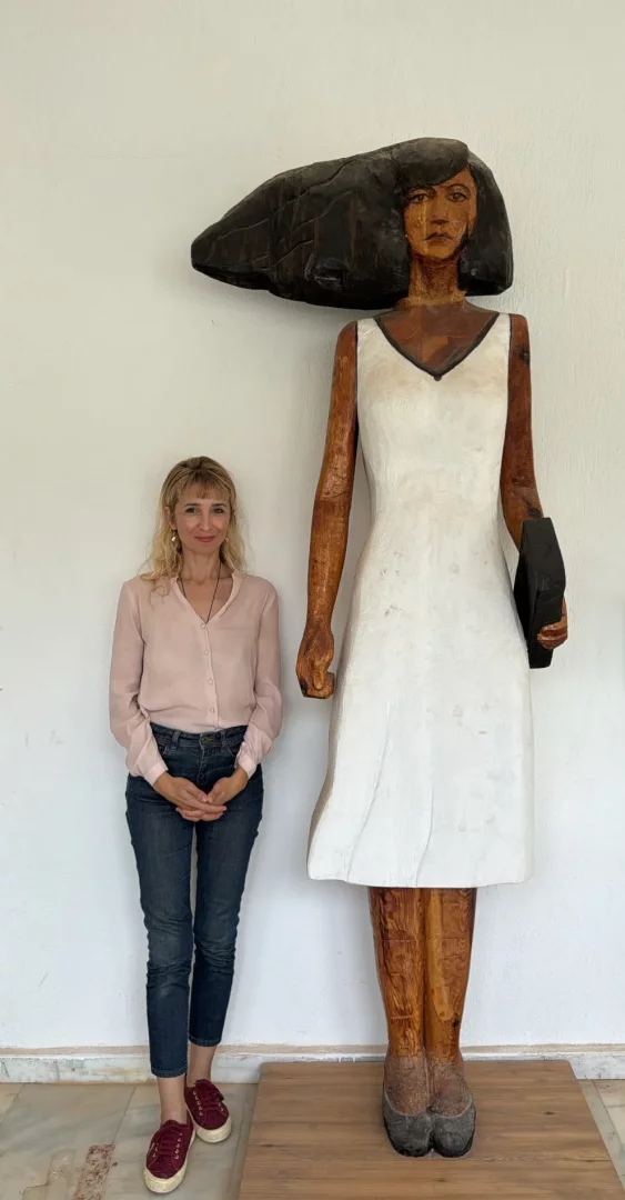 Beyaz bir duvar önünde, sarı saçlı, pembe bluzlu bir kadın, Bodrum atölye buluşmaları etkinliğinde sergilenen, kendisinden kat kat uzun, koyu renk ahşap tonlarında ten rengine ve bembeyaz bir elbiseye sahip, elinde koyu renk bir kitap tutan, etkileyici bir kadın heykelinin yanında poz veriyor.