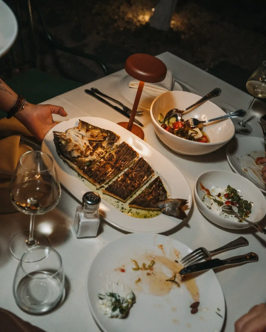 Gece karanlığında bir restoran masasında, ortasında iştah açıcı şekilde dilimlenmiş, yeşil soslu bütün bir ızgara balık tabağı, yanında salata, peynir ve hafif yemek artıkları olan tabaklar, bir tuzluk, kadehler ve loş ışık veren şık bir masa lambası görülüyor.