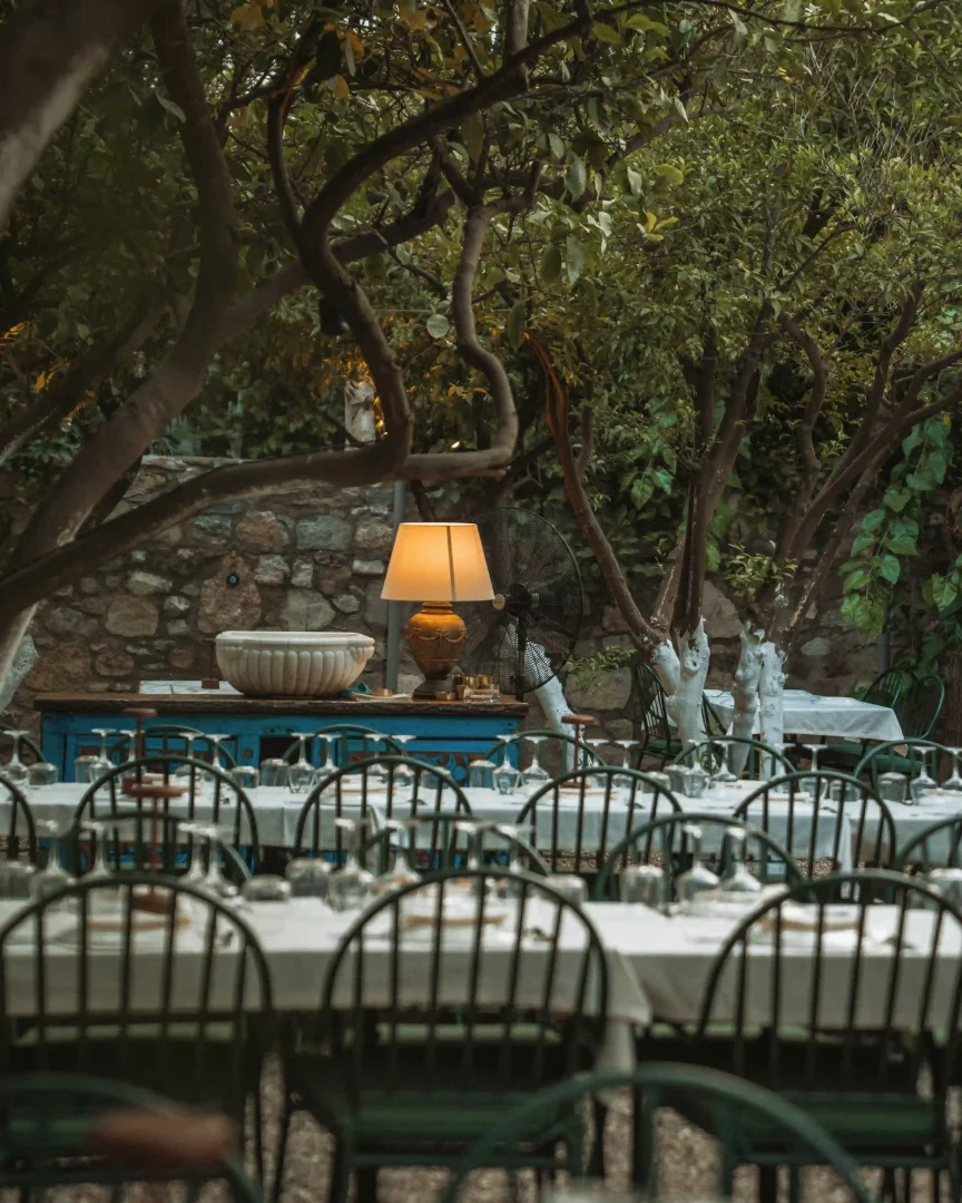 Bir bahçe restoranının akşamüstü atmosferi, beyaz örtülü ve kadehlerle donatılmış çok sayıda masası, yemyeşil ağaçların gölgesinde, taş duvara yaslanmış rustik mavi konsol masası ve sıcak ışıklı lambasıyla huzurlu bir yemek deneyimi sunuyor.