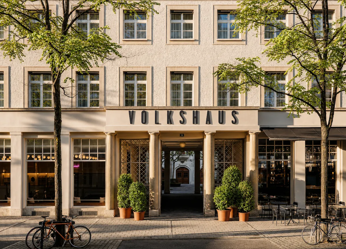 Volkshaus Basel butik tasarım otelinin dış cephesi, önünde genç yapraklı ağaçlar, iki bisiklet, saksı bitkileri ve açık hava oturma alanı bulunan, sıcak bir gün ışığında aydınlanan klasik bir mimariye sahip binayı gösteriyor.