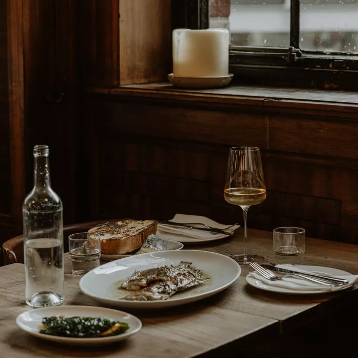 Brat London restoranının otantik atmosferinde, loş ışıklı ahşap bir masada servis edilmiş, büyük oval bir tabakta ana yemek olarak görünen bütün ızgara balık, yanında yeşil sebzeler, kızarmış ekmek, bir kadeh beyaz şarap ve su bulunan davetkar bir yemek düzenlemesi.