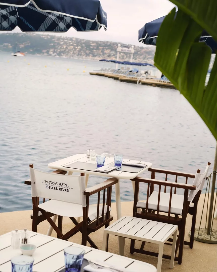 Fransız Rivierası'ndaki Hôtel Belles Rives'in deniz kenarındaki Burberry beach club iş birliğinde, markanın adının yazılı olduğu beyaz bir sandalye, mavi ekose desenli şemsiyeler altında düzenlenmiş beyaz masa ve sandalyeler, mavi bardaklar ve uzakta yüzen bir iskele görülerek moda estetiği sergileniyor.
