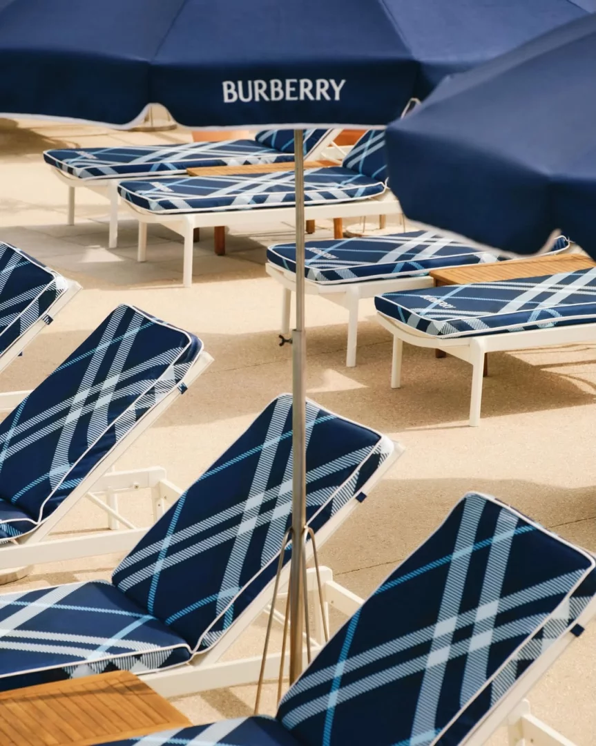 Görselde, Fransız Rivierası'ndaki Hôtel Belles Rives'in Burberry'nin ikonikleşmiş mavi ekose deseniyle kaplı şezlongları ve logosunu taşıyan şemsiyeleriyle tasarlanmış beach club alanı, 2026 yazının öne çıkan moda beach club iş birliklerinden biri olarak lüks tatil deneyimini temsil ediyor.