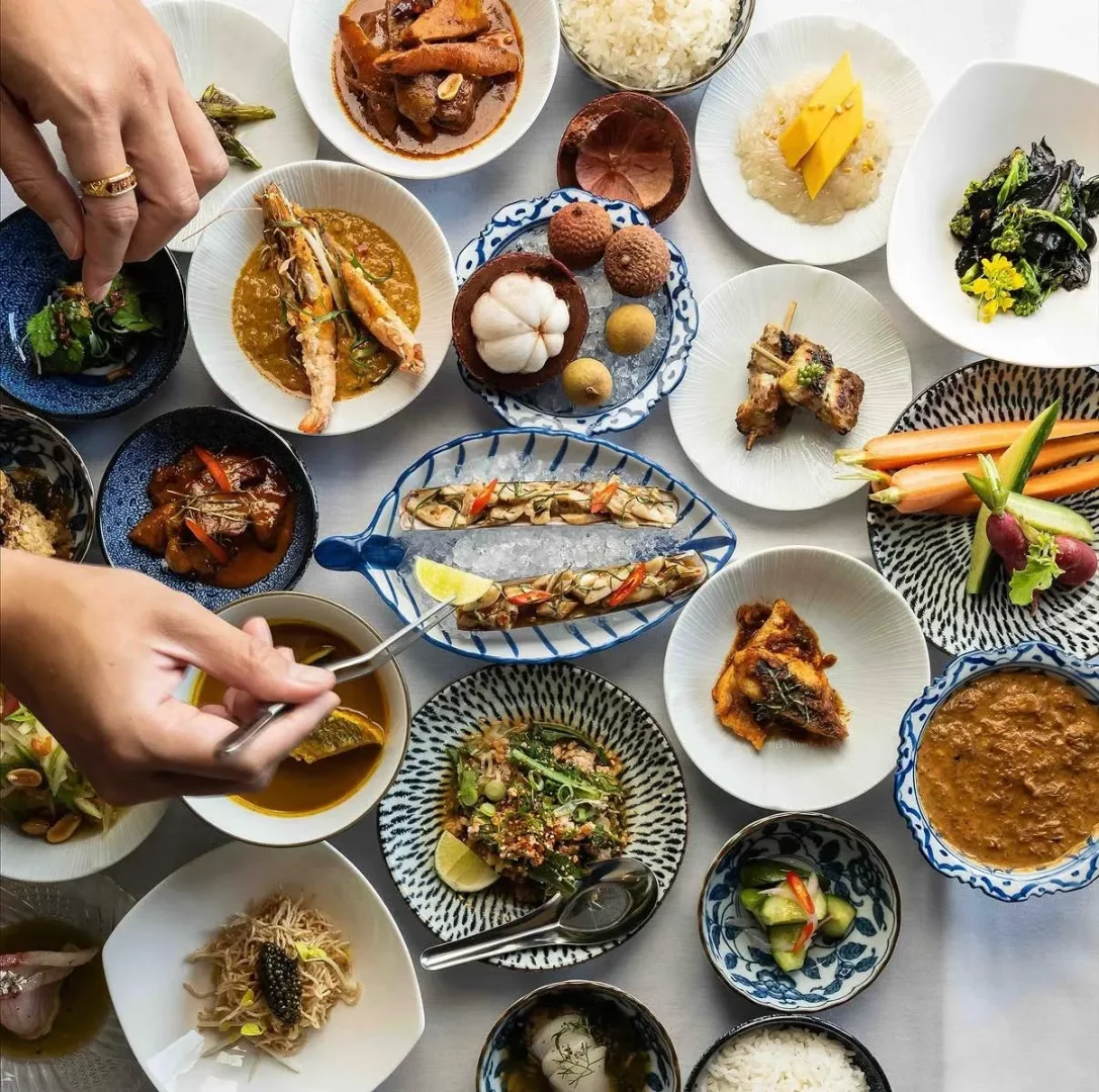 Üstten çekilmiş bir fotoğrafta, beyaz bir masanın üzerinde özenle düzenlenmiş, karidesli köri, mango yapışkan pirinci, satay şişler, taze sebzeler, jilet istiridyeler, mangosteen ve liçi gibi egzotik meyveler de dahil olmak üzere çok sayıda Tayland yemeği ve tatlısının sergilendiği zengin bir sofra görülüyor; iki el yemekleri servis ediyor veya tadım yapıyor.