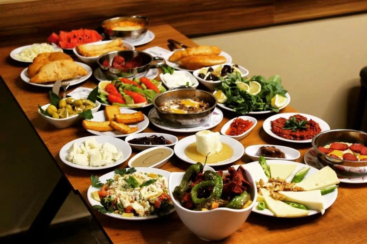 Ahşap bir masa üzerinde, menemen, sucuklu yumurta, beyaz peynir çeşitleri, yeşil zeytin, bal kaymak, tahin pekmez, kızarmış hamur işleri, salata ve karpuz dilimleri gibi geleneksel lezzetlerin bulunduğu, adeta görsel bir şölen sunan zengin bir Türk serpme kahvaltı sofrası.