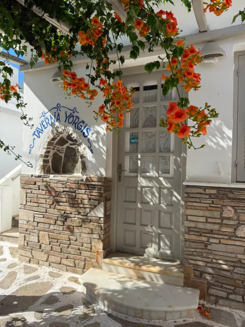 Paros'ta, beyaz badanalı bir binanın taş detaylı girişinde, kapının üzerini saran parlak turuncu trompet çiçekleri ve mavi yazıyla süslenmiş Taverna Yorgis tabelası güneşin altında canlı bir görünüm sunuyor.