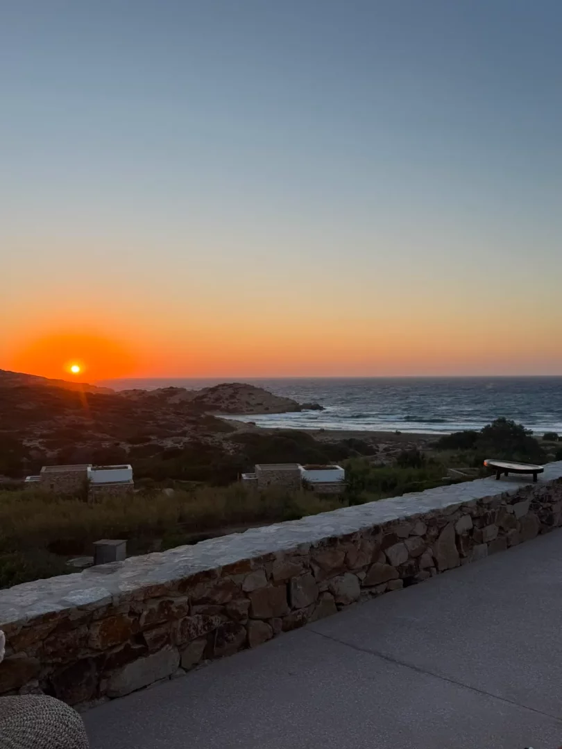 Gün batımında Ege Denizi'nin turuncu ve mavi tonlarındaki gökyüzü altında dalgalı suları, Paros adasının kayalık kıyılarındaki modern taş evleri ve yeşillikleri gözler önüne seriliyor.
