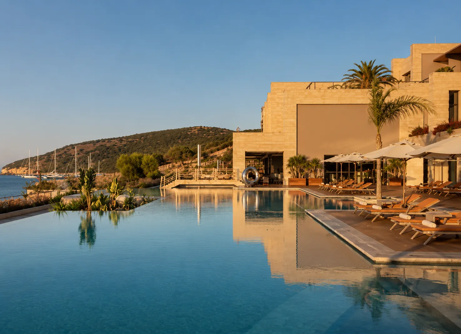 Sonsuzluk havuzunun berrak mavi suları ön planda, Caress Luxury Collection Bodrum Resort'un şık taş binası, havuz kenarında sıralanmış şezlonglar ve beyaz şemsiyelerle lüks bir dinlenme alanı oluştururken, arka planda Ege Denizi'nde demirli yelkenliler ve yemyeşil tepeler gün batımının sıcak tonlarıyla huzurlu bir tatil ortamı yansıtıyor.