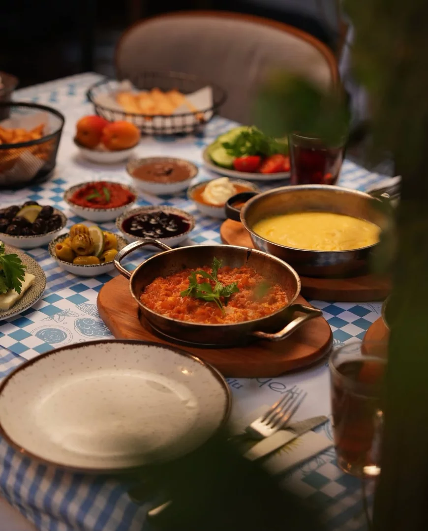 Mavi beyaz kareli masa örtüsü üzerinde, menemen ve mıhlama gibi sıcak lezzetlerin yanı sıra farklı zeytinler, reçeller, taze sebzeler, patates kızartması ve çay bardaklarıyla donatılmış zengin ve geleneksel bir Türk kahvaltısı sofrası görseli.