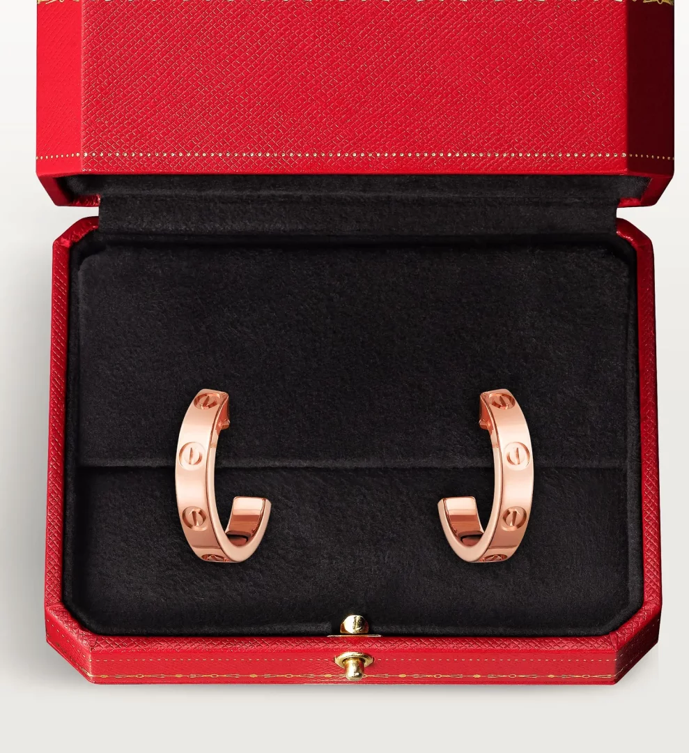 Kırmızı renkli, açılmış lüks kutusunun siyah kadife iç kısmında özenle yerleştirilmiş, Cartier'in ikonik Love koleksiyonuna ait, vida motifleriyle bezeli, iki adet parlak gül altın rengi halka küpe, şık ve zarif bir hediye sunumunu gözler önüne seriyor.