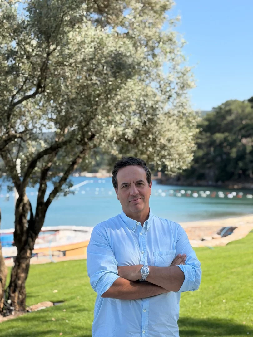 Cem Mira, açık mavi gömleği ve kolları bağlı duruşuyla, arkasında berrak mavi denizin, yeşil çimenlerin ve zeytin ağacının yer aldığı, güneşli Lucca Beach'in doğal güzellikleri arasında gülümseyerek poz veriyor.