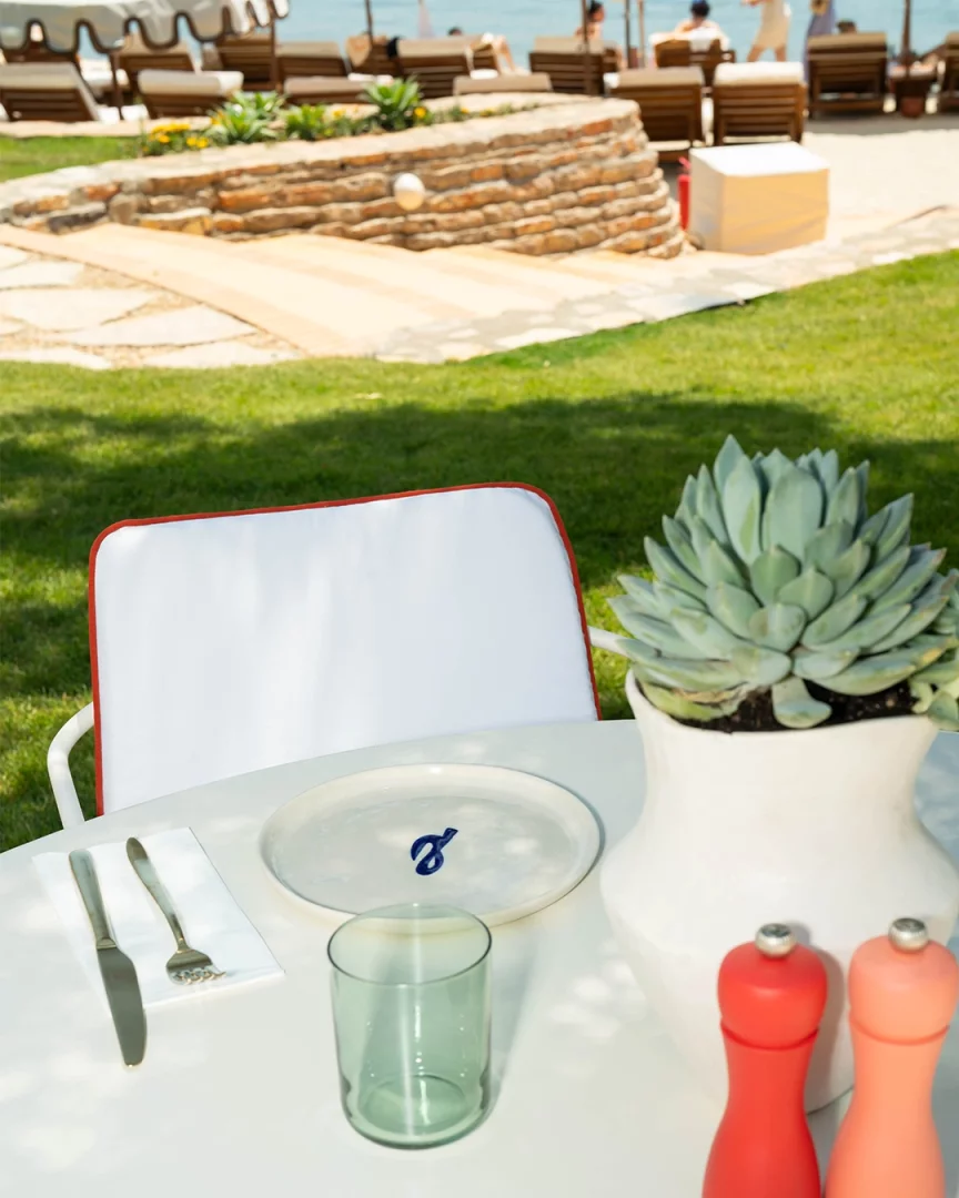 Lucca Beach'in Demirbükü'ndeki yeni döneminde, çimlerin üzerinde özenle düzenlenmiş, minimal tabak takımının ve modern bitki saksısının olduğu bir masada, arka planda denizin ve şezlongların huzur veren manzarasıyla, doğa ve gastronomi odaklı deneyimler sunuluyor.