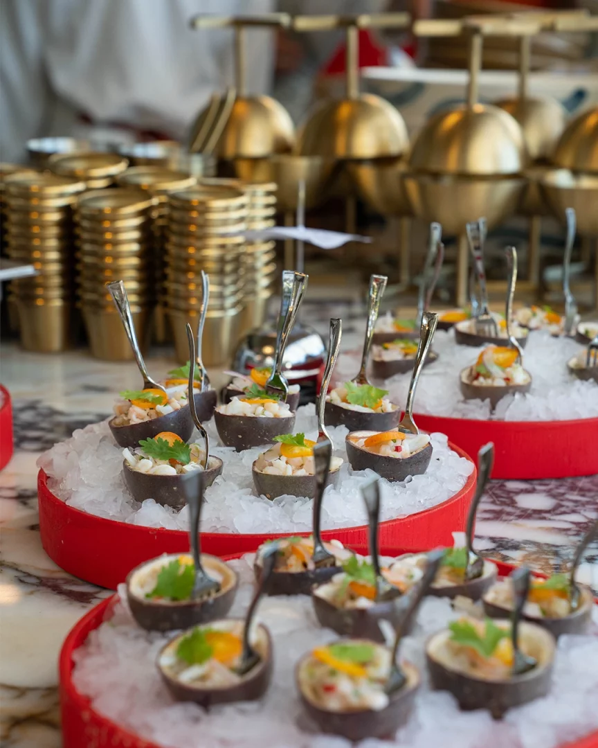 Lucca Beach'in yeni dönemindeki Akdeniz odaklı gastronomi anlayışını sergileyen bu görselde, kırmızı tepsilerde bol buz üzerinde şık bir düzende sunulan, taze deniz ürünlerinden hazırlanmış, yeşillik ve narenciye dilimleriyle süslenmiş onlarca zarif ceviche ve meze, arkadaki altın rengi servis ekipmanlarıyla sofistike bir ikram atmosferi yaratıyor.