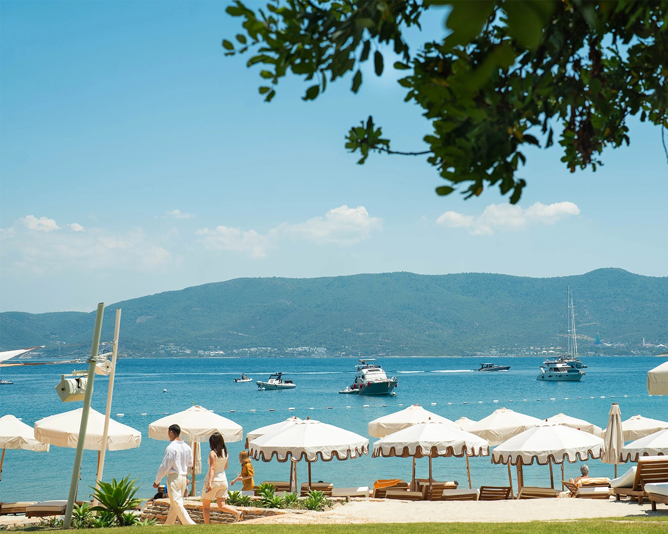 Bodrum'un Demirbükü koyunda konumlanan Lucca Beach'in yeni dönemini anlatan bu karede, bembeyaz şemsiyelerin sıralandığı kumsalda sakin adımlarla ilerleyen şık bir çift ve masmavi denizde seyreden teknelerle Akdeniz yaşam kültürünün huzurlu atmosferi hissediliyor.
