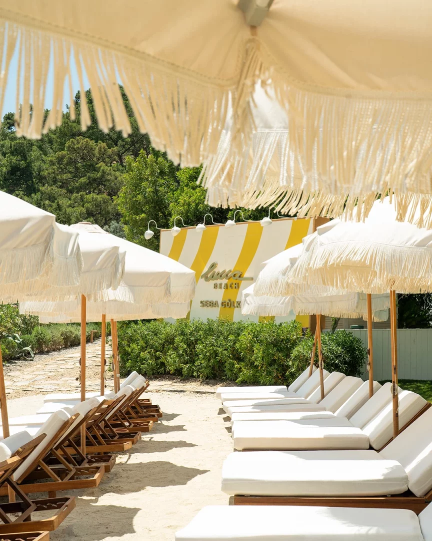 Demirbükü'ndeki Lucca Beach'in yeni dönemini yansıtan bir görselde, beyaz minderli ahşap şezlonglar ve püsküllü şemsiyeler güneşli bir kumsalda düzenli sıralar halinde uzanıyor; arkada Lucca Beach logosunun yer aldığı sarı-beyaz çizgili duvar ve yemyeşil ağaçlar Akdeniz yaşam kültürünün lüks ve huzurlu bir örneğini sunuyor.