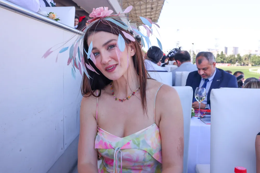 Ceren Benderlioğlu, Gazi Koşusu etkinliğinde pembe ve mavi tonlarda tüyleri olan şık bir şapka, renkli boncuklu kolye ve çiçek desenli askılı elbisesiyle kameralara hafifçe gülümserken, arka planda diğer konuklar ve yarış pisti görünüyor.