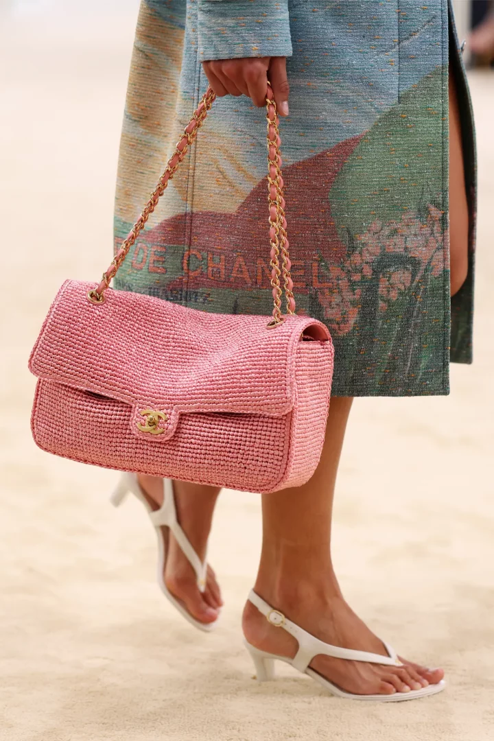 Chanel Cruise 2027 defilesinde, pembe hasır dokulu zincir askılı çantası ve zarif, minimalist beyaz ince bantlı parmak arası alçak topuklu sandaletleriyle podyumda yürüyen bir modelin stil detayı, 2026 İlkbahar-Yaz sandalet modellerinin çıplak ayak estetiğini vurguluyor.
