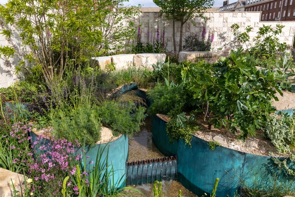Chelsea Flower Show 2026'nın öne çıkan bahçe trendlerinden biri olan suyun estetik ve sürdürülebilir kullanımını vurgulayan bu tasarım, bakır oksitlenmesini andıran metal kenarlarla çevrili akışkan bir su kanalı, çevresinde ise mor süs bitkileri ve gür yeşil çalılıklarla doğal ve kontrollü bir vahşilik sergiliyor.