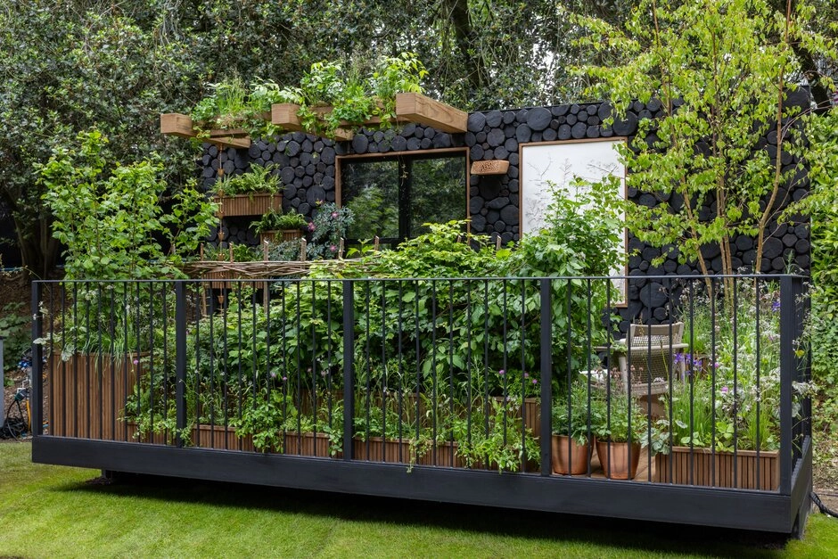 Chelsea Flower Show'un 2026 bahçe trendleri arasında dikkat çeken; siyah kütük uçlarından oluşan modern bir duvarın önünde, bol yeşillik ve farklı bitki türleriyle dolu saksılarla çevrili, kentsel alanları yaşanabilir ekosistemlere dönüştüren doğal ve sürdürülebilir bir balkon veya küçük bahçe tasarımı.