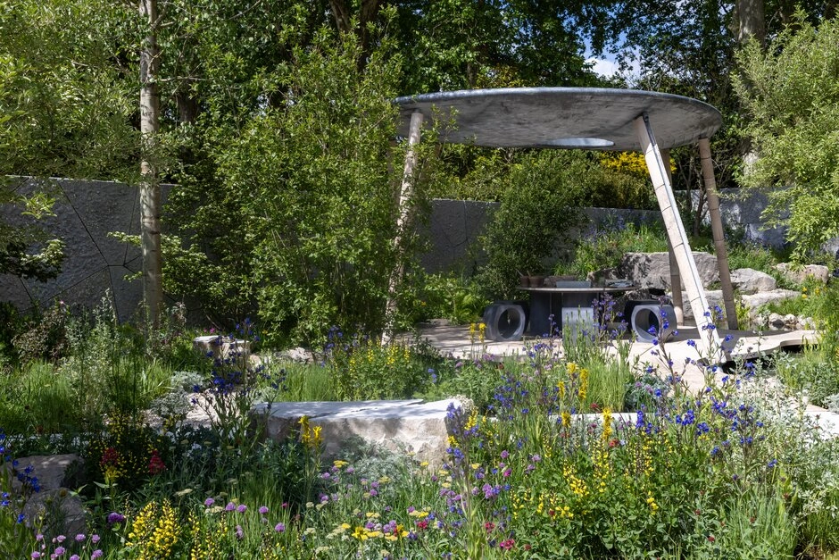 Kontrollü vahşilik temalı bir Chelsea Flower Show bahçe trendi örneği olan bu geniş peyzaj, yer yer irili ufaklı taşlarla bezenmiş doğal bitki örtüsü içinde, canlı sarı, mor ve mavi kır çiçekleriyle dolu; arka planda ise modern, düz gri bir gölgelik ve etrafı yeşil ağaçlarla çevrili gri dokulu bir duvar görülüyor.