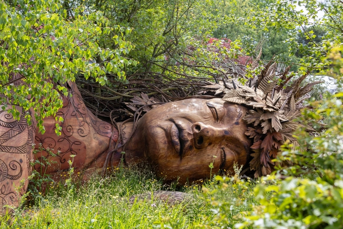 Chelsea Flower Show 2026'da Yılın Bahçesi seçilen Sarah Eberle'nin 'On the Edge' tasarımından, düşmüş ağaçlardan oyulmuş, başı yaprak ve dallarla bezenmiş, otların arasına huzurla uzanmış Toprak Ana heykeli, kontrollü vahşilik ve doğal bahçe trendlerini yansıtıyor.
