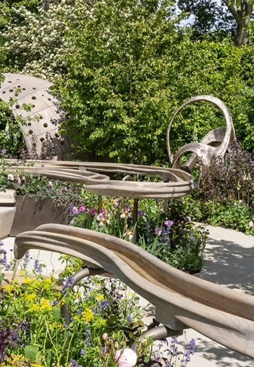 Ön planda su akıtan iki kıvrımlı ahşap kanalın yemyeşil bitki örtüsü ve rengarenk çiçeklerle çevrili olduğu bu Chelsea Flower Show 2026 bahçe tasarımı, doğal dokular ve çağdaş su kullanımı gibi yenilikçi bahçe trendlerini sergiliyor.