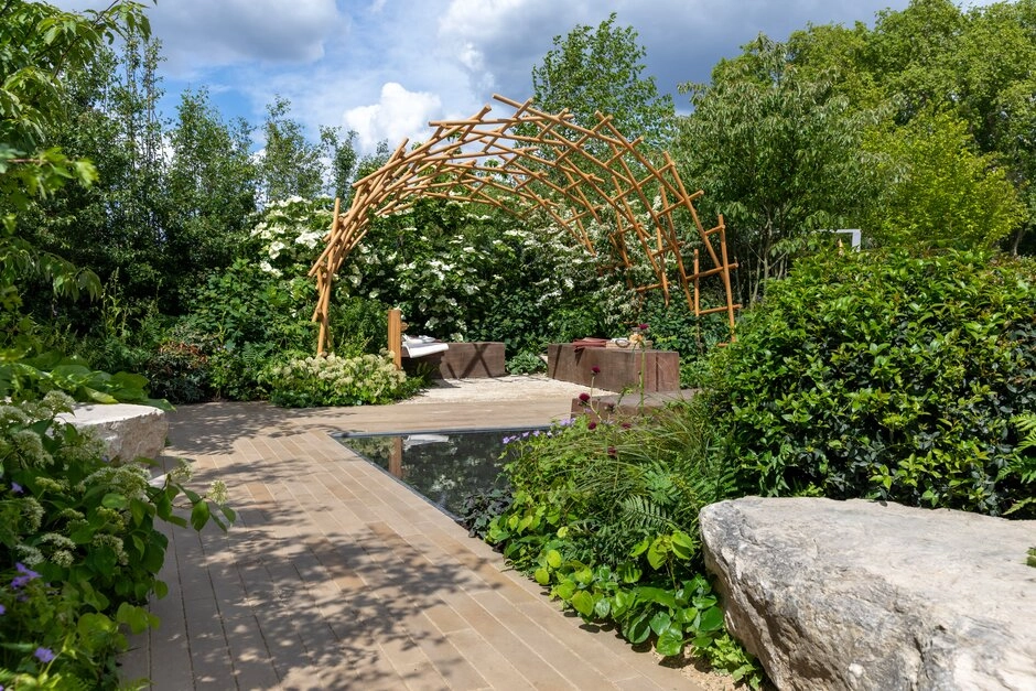 Chelsea Flower Show'un 2026 bahçe trendlerini yansıtan bu tasarımda, doğal ahşap dallardan oluşan organik kemerli bir yapı, dingin bir su havuzu ve yemyeşil bitki örtüsüyle çevrelenmiş, doğayla uyumlu, sürdürülebilir ve huzurlu bir dinlenme köşesi sunuluyor.